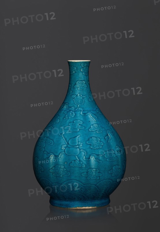 Vase in the shape of an onion-shaped bottle with a narrow, short neck. Onion-shaped bottle-vase decorated in flat relief with a composition combining cloud and babao emblems eight precious objects. Single-color dark green-blue glaze. Ceramics, Chinese, Qing 1644–1911 style; possibly 16th–17th century. Height 27 cm; weight 2.6 kg., A teardrop-shaped ceramic vase with a narrow neck and flared rim sits against a dark gray background, finished in a rich turquoise-blue glaze with a matte, slightly crackled surface; the body is decorated with a repeating raised pattern of stylized cloud-like motifs and flowing ribbon-like lines, with a band of wave patterns circling the lower portion, and a thin unglazed or worn ring of a light beige color visible at the base and rim.