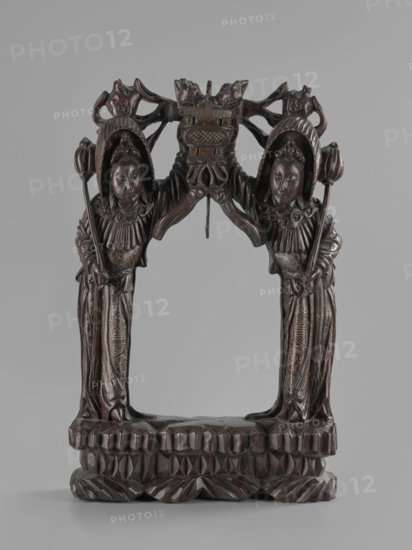 Stand, Chinese, Chinese art, figurative motifs, women, Qing 1644–1911, stand for hanging a gong, 19th century, China, varia, height 45.5 cm, width 29.5 cm, A carved wooden sculpture depicting a symmetrical archway supported by two standing figures facing forward, each wearing ornate headdresses and long robes with carved patterns, holding a long staff topped with a rounded element; the figures have rounded faces with calm expressions and detailed facial features. Above them, carved ornamental elements form a curved top with stylized animal and floral shapes and a central suspended element hanging down from the arch. The base is a carved platform with geometric and leaf-like patterns. The visible colors are dark brown wood tones with subtle lighter brown highlights and faint areas of ochre or tan in carved detailing, set against a neutral light gray background.
