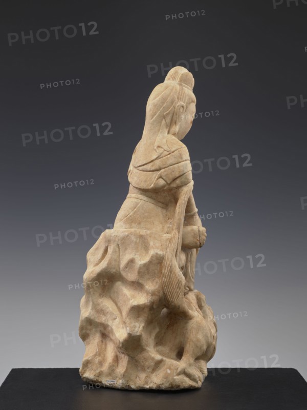 Guanyin with qilin, Chinese, Qing dynasty 1644–1911, 19th century, China, Chinese Buddhism iconography, goddesses, unicorns qilin, sculpture, figurine, height 47.5 cm, A carved stone figure in beige and light tan tones sits on an irregular textured base of the same material, shown from a rear three-quarter view; the figure has long hair pulled into a top knot and a long braided or ribbon-like strand falling down the back, layered clothing with folded drapery across the shoulders and back, and hands held near the front of the body; the stone surface shows smooth and carved grooves, with variations of beige, tan, and hints of brown, set against a neutral dark gray to charcoal background and resting on a flat black platform.