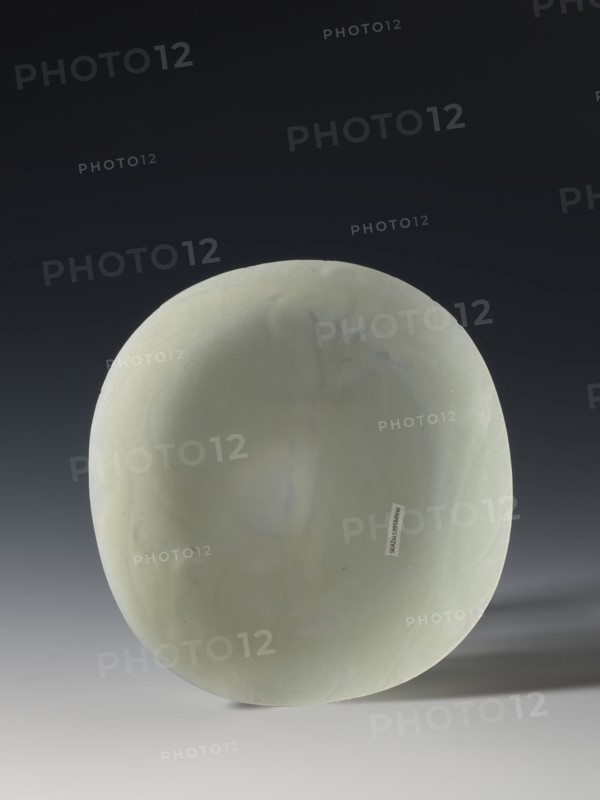 Plaque-lid, 19th century Qing dynasty 1644–1911, China; bas-relief carved jade with engraving; decoration of bats and plant motifs; three double recesses on the bottom; height 14.2 cm, width 15.4 cm., A smooth, rounded object with a pale green surface sits against a dark to light gradient background, showing subtle variations in green and faint bluish-gray veining; the object casts a soft shadow to the right and rests on a light gray surface, and the overall scene includes shades of dark gray, black, pale green, bluish-gray, and light gray.
