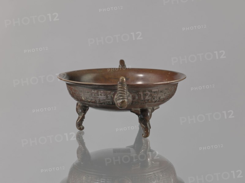Ding vessel, gift, Japanese art, Japanese culture, tripod, Edo 1603–1868, bronze, Meiji 1868–1912. Ding vessel with three legs and two handles. Used for ritual hand washing. 19th century Edo/Meiji, Japan. Vessel; height 9.1 cm, width 24.2 cm, diameter 19.6 cm., A shallow round bowl with ornate relief decoration sits on three sculpted legs, each leg curving and ending in a rounded foot, and the bowl has a small raised handle-like projection on its rim; the object shows a metallic surface with brown and bronze tones and areas of darker patina, and it is reflected on a smooth gray surface beneath; the background is a uniform light gray.