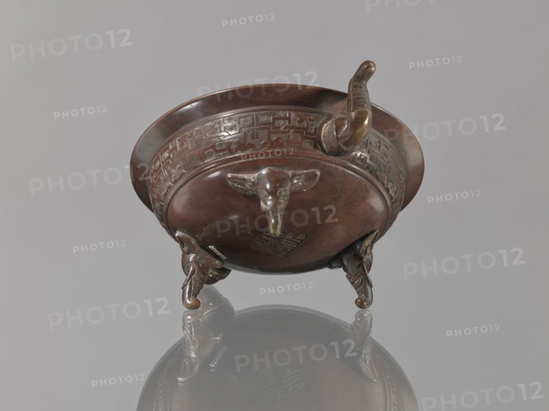 Ding vessel, Ding vessel with three legs and two handles. Used for ritual hand washing. Bronze. Japanese; Edo 1603–1868 / Meiji 1868–1912, 19th century. Height 9.1 cm; width 24.2 cm; diameter 19.6 cm., A bronze vessel with a round body and wide rim sits on a reflective grey surface, showing a polished brown and dark brown patina with greenish highlights in places; the vessel has decorative raised banding around the upper body with geometric motifs, a protruding curved spout or handle, and three sculpted animal-head feet each with curled details; the reflection of the vessel is visible below, and the background is a uniform light grey.
