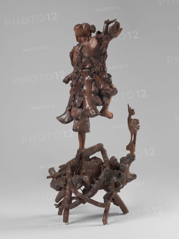 Dancing with a bird, unknown sculptor, 19th century, China; sculpture, height 77 cm; subject: dance with a bird; motifs: birds, cranes, figurative motifs; culture: Chinese/Asian; iconography: dance., A carved wood sculpture with a rich brown patina standing on a base of intertwined, irregular wooden limbs and branches, supporting a vertical central figure composed of layered, organic, root-like forms that suggest a cloak or mass around a humanoid torso and head, with a smooth, rounded head form and a bun-like shape at the back; the figure has an outstretched arm and a bent leg emerging from the carved mass, and a smaller carved animal-like figure in a similar brown color stands on a projecting wooden support beside the central composition, all set against a plain light gray background.