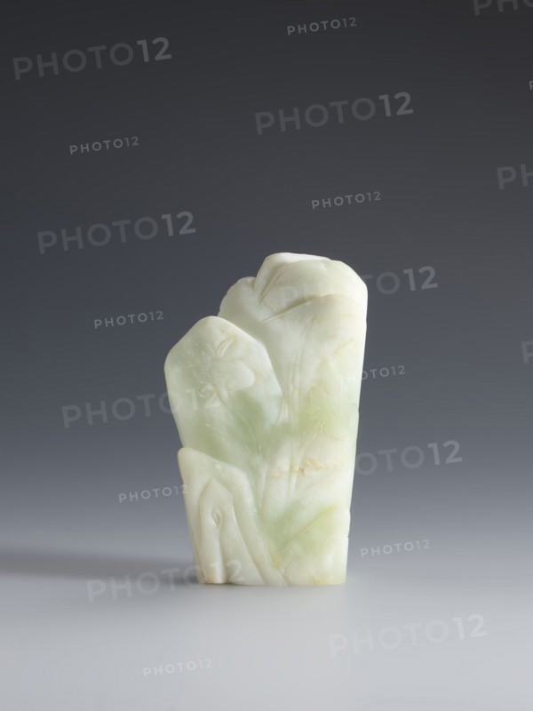 Small screen on a base, Chinese, Qing 1644–1911. Pale green jadeite small screen with an oval cross-section. Engraved decoration: composition of pine, grass, lingzhi mushrooms on rocks; at the top a crescent with the inscription yue. Wooden elongated base, tapering on the shorter side, on four short legs one broken, bulging side inlaid with silver wire. 18th–19th century, China. Sculpture, screen. Dimensions: sculpture height 14.3 cm, width 9.1 cm; base height 8.7 cm, width 4.7 cm., A pale green and white carved stone object stands upright on a smooth gray surface against a darker gray background, showing a layered, rounded top and carved flowing lines and shallow reliefs suggesting organic shapes; the stone displays translucent areas, subtle pale yellow veins, and smooth polished surfaces with softer matte patches, casting a soft shadow to one side.