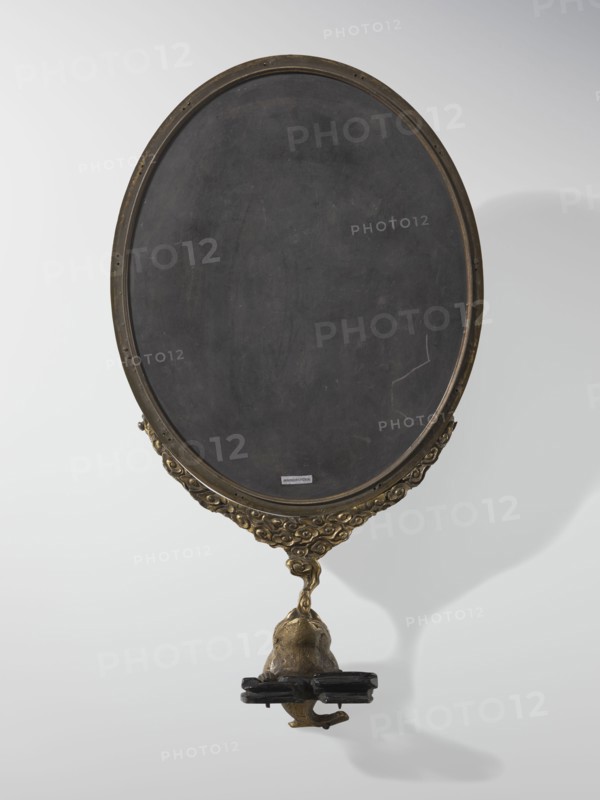 Framed oval screen, Chinese, Qing 1644–1911. Oval screen made of black slate in a gilded bronze frame topped with a frog figurine; depicts children and children's games. 19th–20th century, China. Height: 42.0 cm., An oval object with a dark gray to black central surface set in a bronze-colored frame showing ornate carved patterns along the lower edge, attached to a bronze-colored sculpted support that tapers into a small figure or form holding a dark brown to black horizontal element, all photographed against a pale gray to off-white background with soft shadows.