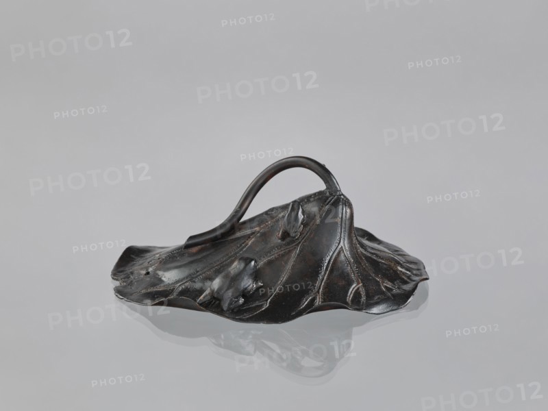 Lotus Leaf with Two Frogs, Ryûbundô,  craftsman author; Japanese; frogs; paperweight; lotus leaf; sculpture; Japan, 2nd half of the 19th century; height 6.7 cm, width 18.2 cm., A dark metallic object shaped like a large, slightly crumpled leaf with raised veins and organic folds sits on a smooth reflective surface, showing a muted reflection beneath it; a curved handle arches from one side to the other, and the surface of the object displays subtle highlights and worn areas in dark brown and black tones against a pale gray background.
