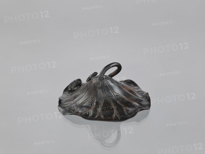 Lotus Leaf with Two Frogs, Ryûbundô,  craftsman author, Japanese culture, Japanese art, frogs, Japanese, 2nd half of the 19th century, Japan, sculpture, paperweight, lotus leaf form, height 6.7 cm, width 18.2 cm, A dark metallic object shaped like a broad, flattened leaf with undulating edges sits on a reflective pale gray surface; the object has visible raised vein-like ridges and a central looped handle rising from its middle, with a few small rounded protrusions near the handle. The surface shows highlights of brownish bronze and black tones, and the reflection of the object appears beneath it on the pale gray background.