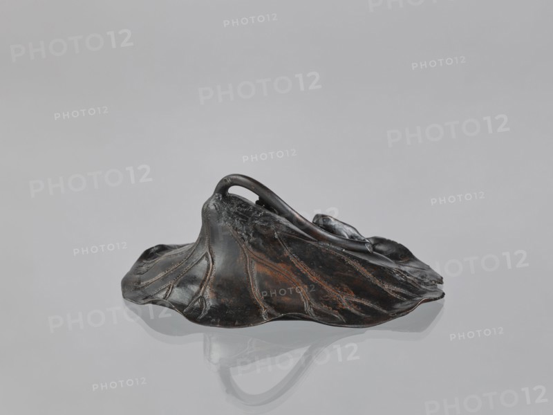 Lotus Leaf with Two Frogs, Ryûbundô,  craftsman author; Japanese; frogs; paperweights; lotuses; leaves; sculpture; Japan, 2nd half of the 19th century; height 6.7 cm, width 18.2 cm, A dark, irregularly shaped sculptural form resembling a wilted leaf or hat sits on a smooth, reflective surface; the object shows a mix of black and brown tones with subtle reddish-brown highlights and worn areas, and it has raised, vein-like ridges and a curved handle-like loop rising from its center, with a soft reflection of the object visible on the pale gray surface beneath.