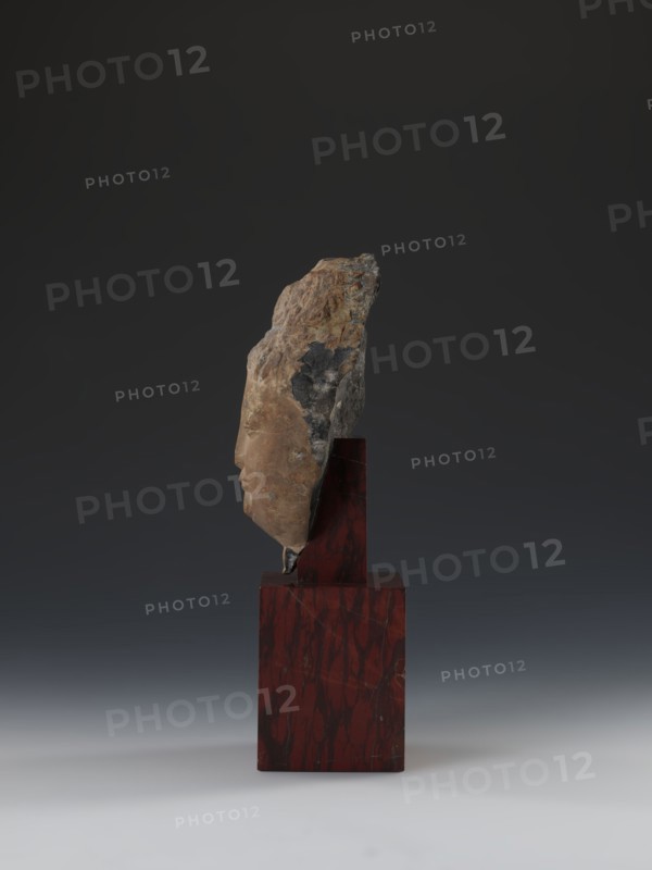 Head of Guanyin Bodhisattva, sculptor unknown. Polychrome-coated feldspar fragment of a statue with a cube-shaped base of dark red marble. Chinese; Qing dynasty 1644–1911, 19th century. Iconography: Guanyin. Sculpture; height 26.0 cm., A fragmentary stone sculpture of a human profile mounted on a polished rectangular red and dark marbled base, the stone showing rough, weathered surfaces with brown, tan, gray, and black tones while the carved facial area is smoother and beige, set against a gradient dark gray to light gray background.