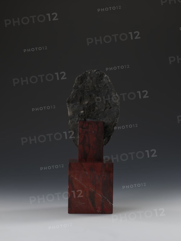 Head of Guanyin Bodhisattva, Chinese, Guanyin iconography, Qing style, polychrome-coated feldspar with cube-shaped base of dark red marble, 19th century Qing dynasty, 1644–1911, China, sculpture, height 26.0 cm, A dark gray, irregularly shaped rock is mounted on a polished red stone pedestal with visible darker veins; the pedestal consists of a larger red block topped by a smaller red column, both showing marbled patterns. The background is a smooth gradient from deep charcoal at the top to a lighter gray near the bottom, and the surface beneath the pedestal is a soft gray transitioning to an off-white foreground. The visible colors are dark gray, black, red, maroon, and shades of gray.