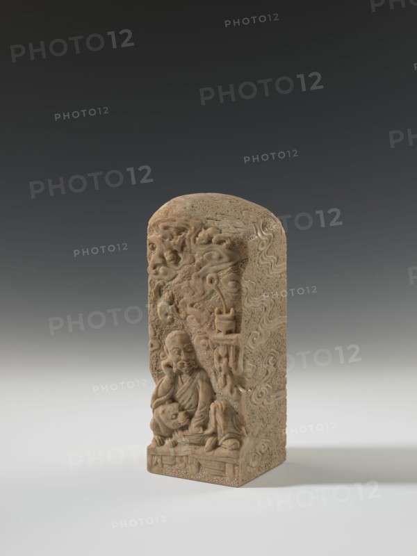 Seal stamp, lion iconography, bats iconography, arhat iconography, Chinese art, seal stamps, Chinese culture, Qing 1644–1911, bas-relief decoration, 19th century, China, seal stamp, height 23.6 cm, width 8.9 cm, depth 9.0 cm, A carved rectangular stone object with a rounded top stands on a flat surface against a gradient background. The object is a beige to light brown color with subtle darker brown variations and textured surface details. The front face features a carved human figure seated in a contemplative pose, surrounded by swirling cloud-like motifs and additional carved elements that project from the surface. The sides and top of the object display carved patterns and textured areas consistent with weathered stone. The lighting casts a soft shadow to the right and the overall scene includes shades of gray in the background and white on the surface beneath the object.