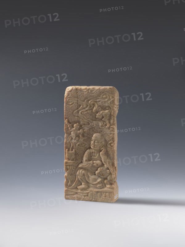 Seal stamp;, bas-relief decoration depicting bats, a lion, and an arhat; Chinese; Qing dynasty 1644–1911, 19th century ; seal stamp in stone; height 20.0 cm, width 9.3 cm, depth 2.9 cm., A rectangular carved stone plaque in shades of beige and tan stands upright against a smooth gradient background of dark gray at the top fading to light gray at the bottom, showing a relief scene with a seated robed figure in the lower center surrounded by flowing cloudlike motifs and other carved details, with subtle variations of brownish-beige on the plaque surface and faint shadowing on the light gray surface beneath it.