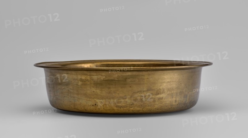 Bowl, round with collar collar decorated with a plant tendril. 19th–20th century, China. Porcelain vessel, height 8.7 cm, diameter 33 cm. Decoration: fruit peaches, pumpkins, pomegranates, lingzhi, images of birds cranes, deer, pine trees, artemisia, plant ornament; Chinese symbols for happiness., A shallow round metal basin with a wide flared rim sits centered against a smooth neutral background; the basin has a warm golden-bronze color with areas of darker patina and subtle greenish and brownish discoloration, visible surface scratches, and small patches of wear; the interior rim shows a slightly brighter golden tone while the exterior surface displays mottled variations in sheen and texture.