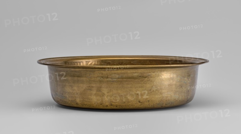 Bowl, fruit, images of birds, bowls, plant ornament, Chinese art, Chinese culture, peaches fruits, lingzhi, pumpkins, pomegranates fruits, happiness symbol, deer, artemisia, cranes, pine trees, Chinese symbols. Round bowl with collar collar decorated with a plant tendril. 19th/20th century, China. Vessel; height 8.7 cm, diameter 33 cm., A shallow, round metal basin with a wide flared rim sits centered against a neutral background; the basin appears made of a warm golden-bronze metal showing areas of darker patina and subtle irregular marks and scratches on its exterior surface, with a slightly reflective inner surface visible along the rim. The background and surface beneath the basin are light gray, and the overall colors visible are gold, bronze, brown, gray, and hints of darker tarnish.