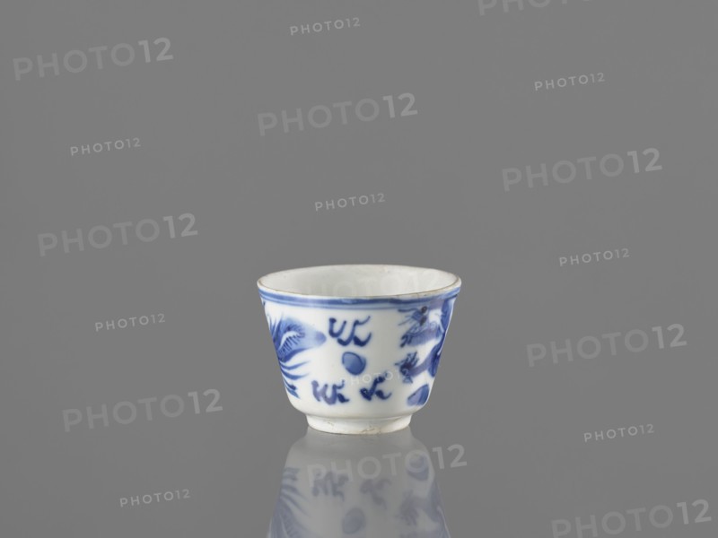 Bowl, Chinese, 19th century. Blue-and-white porcelain vessel decorated with dragons, phoenixes fenghuang, and birds; pearls motif. Height 4.6 cm, diameter 5.9 cm., A small ceramic cup with a white base and blue painted designs sits on a reflective surface, showing a faint mirrored reflection below; the cup has a slightly flared rim with a thin blue band, blue brushstroke motifs around the exterior that resemble abstract shapes and floral elements, and a smooth white interior; the background and surface are shades of gray.