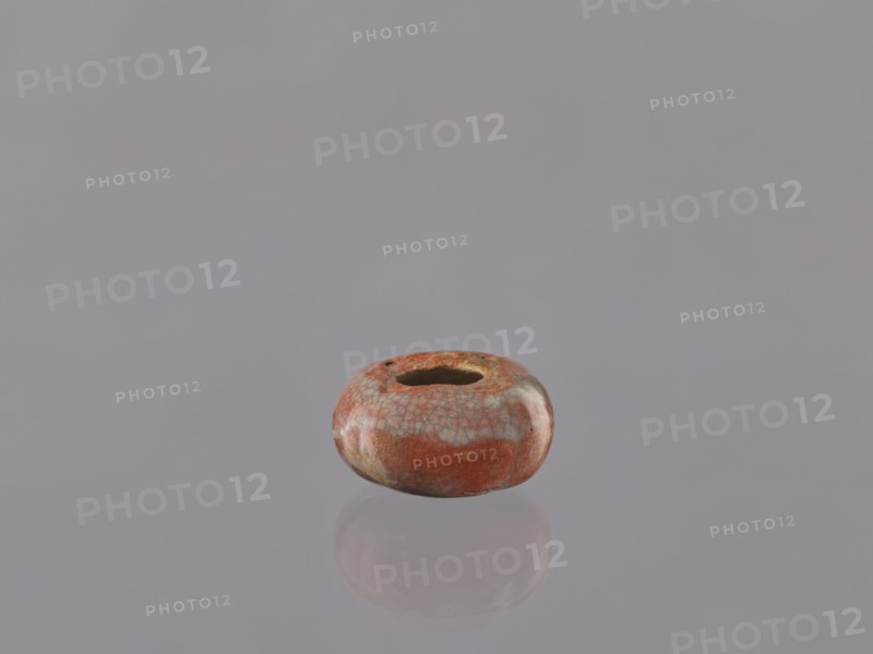 Cosmetic vessel, hand-made vessel head made of yellowish clay, hand-formed. Reddish glaze with gray and blackish streaks, with cracklework. Rusty vessel with dark and light streaks. Probably missing lid. 19th century, Japan. Vessel, height 3 cm, diameter 6 cm. Materials/technique: glazed ceramics, cracklework. Classification: Japanese ceramics., A small rounded object with a central hole sits on a smooth reflective surface, showing a soft reflection beneath it; the object has a glossy finish with a mottled pattern in shades of reddish-brown and muted orange, with patches of pale gray and subtle greenish-beige, and the surrounding background and surface are a uniform light gray.