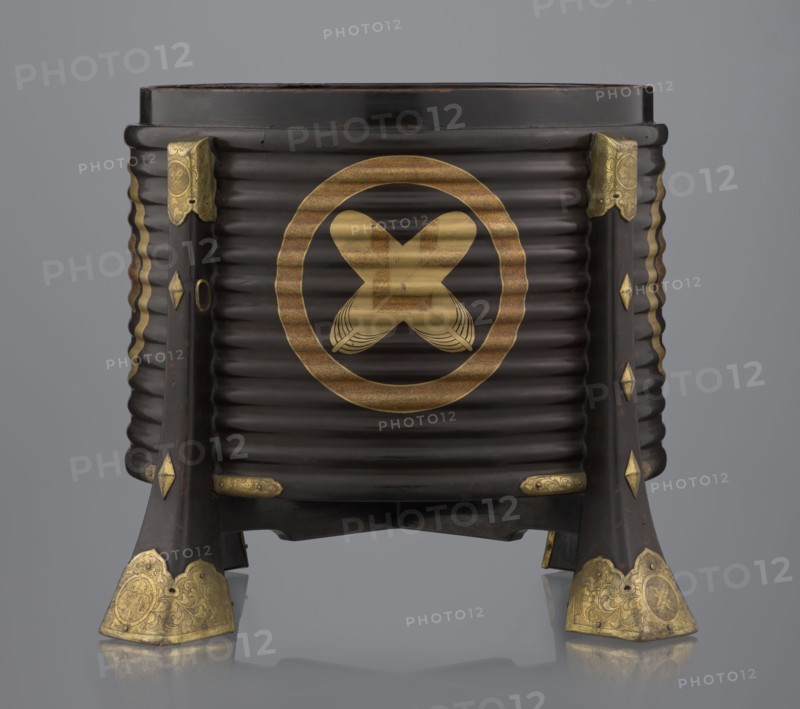 Container hokai for shells for the game kai awase;, Japanese; Meiji 1868–1912 and Edo 1603–1868 periods; wood covered with black lacquer, decoration with gold maki-e lacquer, gilded copper fittings; decorated with feathers and aoi hollyhock plant ornament; boxes and shells for parlor/game use; 19th century Edo–Meiji; place of origin: Edo, Japan; container; height 37.5 cm, diameter 40.0 cm., A dark brown cylindrical wooden container with horizontal ridges stands on four flared legs; the container features several gold-colored metal fittings and ornamentation at the top corners, along the legs, and on the foot ends, and a large circular gold-colored decorative motif is visible on the front; the legs have diamond-shaped gold-colored accents and the foot caps have engraved patterns; the background is a smooth gray gradient and the surface beneath reflects the object.