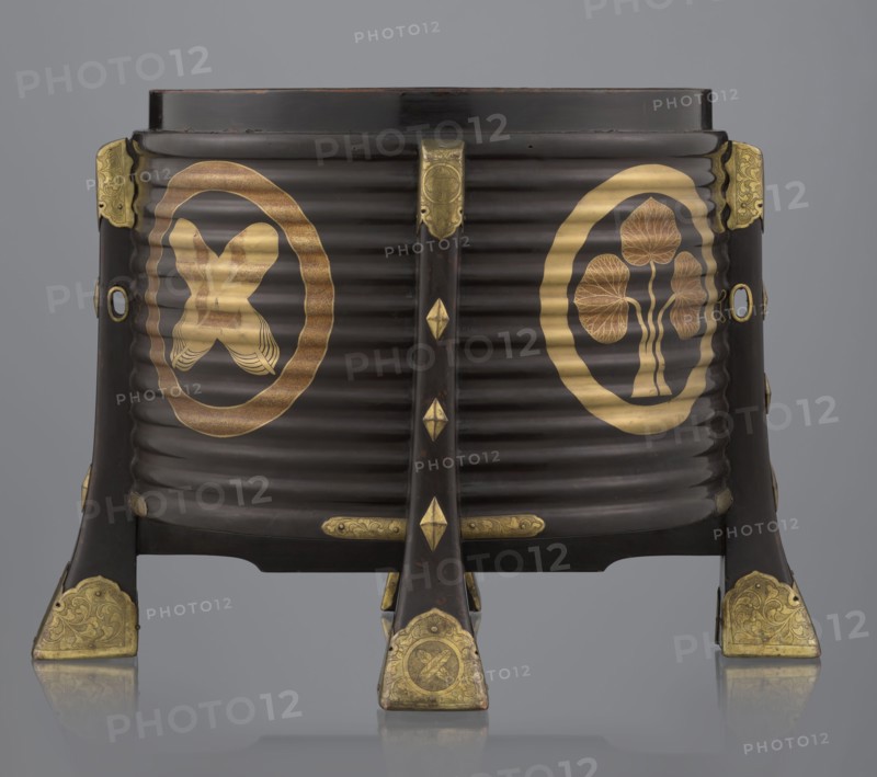 Container hokai for shells for the game kai awase, Japanese, Meiji 1868–1912 and late Edo 1603–1868 periods, wood covered with black lacquer, decoration with gold maki-e lacquer, gilded copper fittings; decorated with feathers and aoi hollyhock plant ornament; boxes, shells, parlor game; 19th century; Edo, Japan; container; height 37.5 cm, diameter 40.0 cm, A dark brown, ribbed cylindrical object set on four angled legs with decorative metal fittings; the surface shows gold circular motifs on the front sides featuring stylized organic shapes and a crossed ribbon-like form, and several gold ornamental plates and diamond-shaped accents on the legs and central support. The visible colors are dark brown, gold, and a neutral gray background.