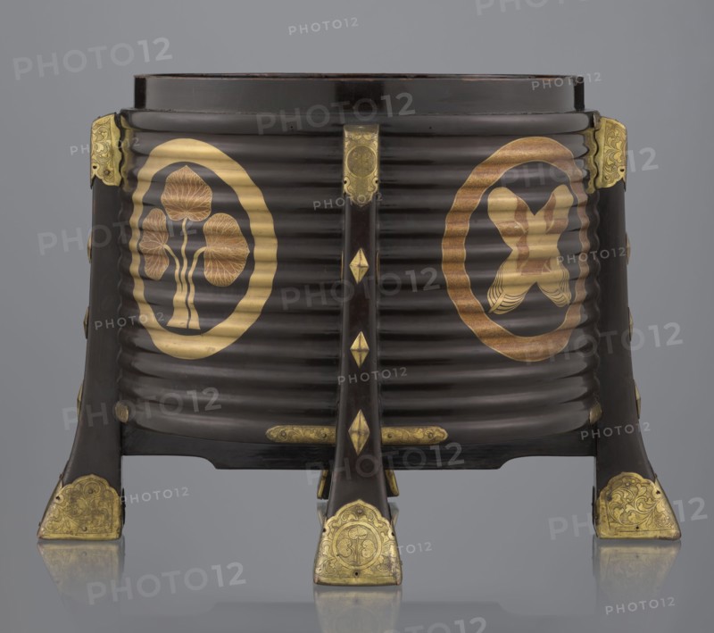 Container hokai for shells for the game kai awase, Japanese, Meiji 1868–1912 and late Edo 1603–1868 periods, wood covered with black lacquer, decoration with gold maki-e lacquer, gilded copper fittings. Decorated with feathers and aoi hollyhock plant ornament. Function: container for shells used in the parlor game kai awase. 19th century Edo–Meiji. Place of origin: Edo, Japan. Height 37.5 cm; diameter 40.0 cm., A dark brown ribbed wooden container with a round top supported by four angled legs, decorated with gold metal fittings on the leg ends and on the top corners; the front shows a pair of circular gold emblems, one with a stylized tree-like motif and the other with an abstract symmetrical motif, both rendered in gold tones with some reddish-gold shading; the legs and central vertical support feature small gold diamond-shaped accents and engraved gold plates with floral patterns; the background is a smooth gray.