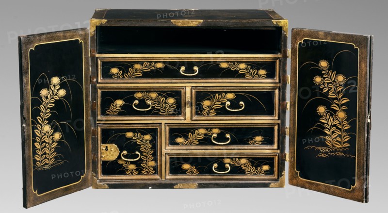 Small cabinet kodansu, Meiji period 2nd half of 19th century, Japan. Rectangular cabinet closed with double doors mounted on engraved brass hinges; equipped with seven drawers the eighth at the top is missing. Door face filled with a landscape composition with pavilions by the water, hills, and a flock of cranes. Side exterior walls decorated with a bamboo motif; interior decorated with floral motifs. Decoration executed in maki-e with gold powder on wood coated with black lacquer. Motifs include flowers, landscape elements, geese, mountains, pines, bamboo, spotted pheasants, and architecture. Piece of furniture; height 45.5 cm, width 52.2 cm, depth 31.7 cm., A rectangular black cabinet with its two doors open to reveal a set of drawers decorated with gold floral motifs; the exterior and drawer faces are glossy black with gold trim and gold handles, and the inner panels of each door feature vertical gold plant designs on a black background framed by a gold border; the drawer fronts dis...