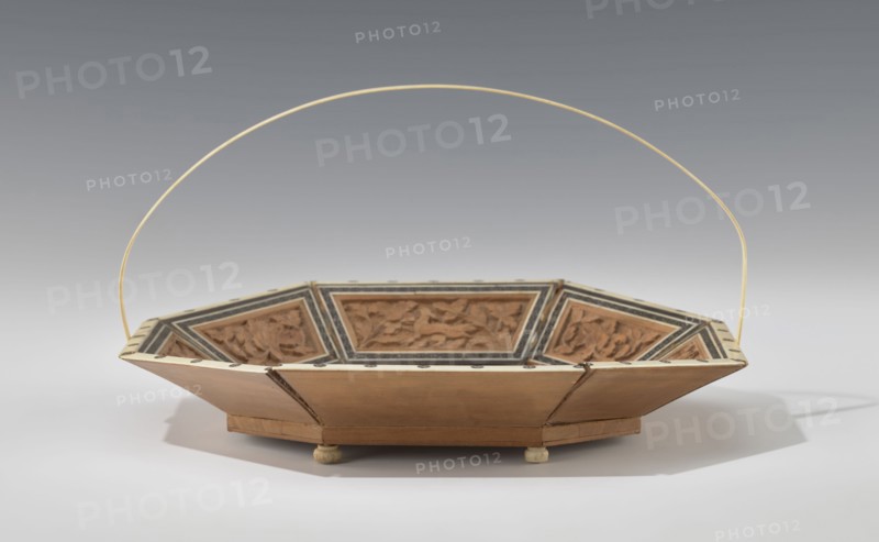 Basket. Octagonal shallow wooden basket with an ivory rim and handle. Inside the basket is a relief scene: in the central field a palace surrounded by rich vegetation, at the foot of which a wild, spotted animal leopard? tiger? dog? strolls. A trail of stars is carved around this scene. The side panels alternate with images of the same animal and flowers; spaces between panels are filled with colorful mosaic tiles. Handle and rim are ivory. Served both utilitarian and decorative purposes. 19th century, India; height 14.0 cm, width 24.0 cm; floral and animal ornamentation., A shallow, octagonal wooden basket with a thin, curved pale yellow handle rising above it, showing carved decorative panels inside the sloping sides; the basket displays natural wood tones, pale beige, light brown, dark brown, and black accents, and rests on small round feet casting soft shadows on a light gray surface beneath a gradient gray background.