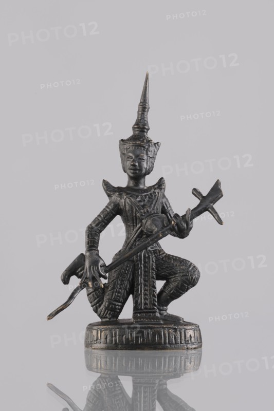 Musician figurine, Southeast Asian art, Southeast Asia culture, musicians, headgear, 1st half of the 20th century, Cambodia place of origin, sculpture, figurine, height 11.1 cm, A small dark metallic figurine of a kneeling figure wearing an ornate pointed headdress and detailed patterned clothing, holding a long curved object across the body; the figure stands on a round decorated base and shows textured surface details and slight worn highlights in a few areas, with colors including dark gray and metallic goldish accents against a pale gray background and a reflective surface below that mirrors the base and parts of the figure.