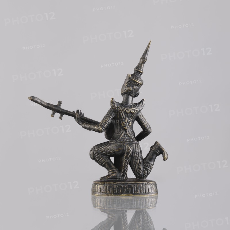 Musician figurine, Southeast Asian art, musicians, Southeast Asia culture, provenance unknown, 1st half of the 20th century contemporary product, Cambodia place of origin, sculpture, figurine, height 11.1 cm, A small dark metallic figurine with worn gold highlights shows a kneeling figure wearing an ornate tall pointed headdress and detailed patterned clothing, holding a long thin staff-like object horizontally with one arm extended; the figure is mounted on a round decorated base and sits against a plain light gray background with a soft reflection of the base visible on the surface below.