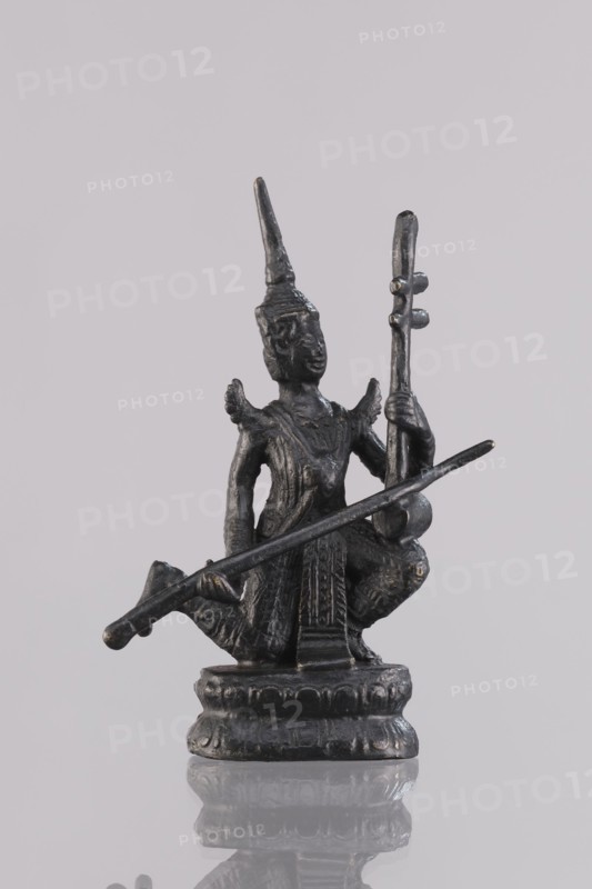 Musician figurine, Southeast Asian art, musicians, Southeast Asia culture, 1st half of the 20th century contemporary product, Cambodia place of origin, sculpture, figurine, height 11.1 cm, A small dark metallic statue of a seated figure wearing a pointed headdress, holding a long thin rod across the body and a taller vertical object in one hand, with decorative ridges and textured clothing, standing on a rounded ornamental base, against a pale gray background; visible colors include dark gray, black, and subtle metallic highlights.