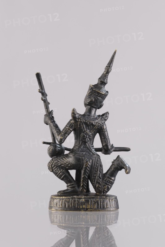 Musician figurine, Southeast Asia culture, art of Southeast Asia, 1st half of the 20th century contemporary product, Cambodia, sculpture, figurine, height 10.9 cm, A small dark metallic statue of a kneeling figure with ornate patterned clothing and a tall pointed headdress, holding a long thin staff in one hand and a rounded object near the other hand; the statue sits on a decorated circular base and shows subtle worn highlights in a gold tone against the primary dark metal color, with a pale neutral background and a faint reflection of the statue visible below.
