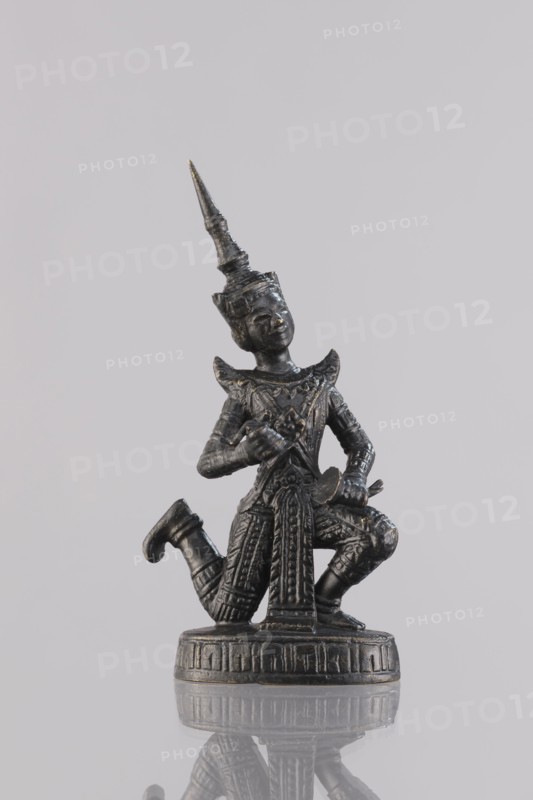 Musician figurine, Southeast Asian art, Southeast Asia culture, musicians, 1st half of the 20th century contemporary product, Cambodia place of origin, sculpture, figurine, height 11 cm, A dark metallic figurine of a kneeling human figure wearing an ornate pointed headdress and intricately patterned clothing is shown against a pale gray background, the figure holding small objects near its chest and lap, standing on a round decorated base with a subtle reflection visible on the glossy surface below; visible colors include dark gray, black, and hints of metallic gold against the light gray backdrop.