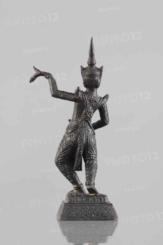 Dancer figurine, Southeast Asian art, musicians, Southeast Asia culture, 1st half of the 20th century contemporary product, Cambodia, figurine, sculpture, height 12.7 cm, A small dark metallic statue viewed from behind showing a standing figure on a decorated rectangular base, the figure wearing an ornate pointed headdress and richly textured clothing with patterned trousers and a draped sash, one arm raised with a curved hand gesture and the other arm bent at the waist, the surface showing highlights of lighter metallic tones against the darker metal, all set against a pale neutral background with a subtle reflection of the base below.