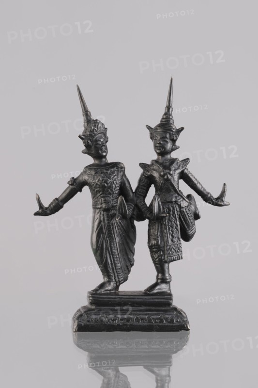 Figurines of a dancing couple, Southeast Asian art, dance, Southeast Asia culture, unknown provenance; figurines of dancers slightly turned to face each other; 1st half of the 20th century contemporary product; Cambodia; figurine, sculpture; height 10.8 cm, A dark metallic statue of two standing figures on a rectangular base is shown against a light grey background; the figures wear tall pointed headdresses and ornate garments with textured patterns, both extending their arms outward with palms facing up, and the surface of the statue reflects subtle highlights of silver and black tones while the base shows worn edges and a faint mirrored reflection on the surface below.