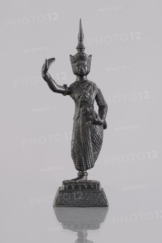 Dancer figurine, Southeast Asian art, Southeast Asia culture, dancers, provenance unknown, 1st half of the 20th century, Cambodia, figurine sculpture, height 12.7 cm, A dark metallic statue of a standing figure is centered against a pale gray background, showing a tall pointed headdress and a detailed textured garment with intricate patterns; one arm is raised with an open hand while the other rests near the waist, and the figure stands on an ornate rectangular base that casts a faint reflection on the smooth surface below. Colors visible include dark gray to black on the statue and various shades of light gray in the background and reflected surface.