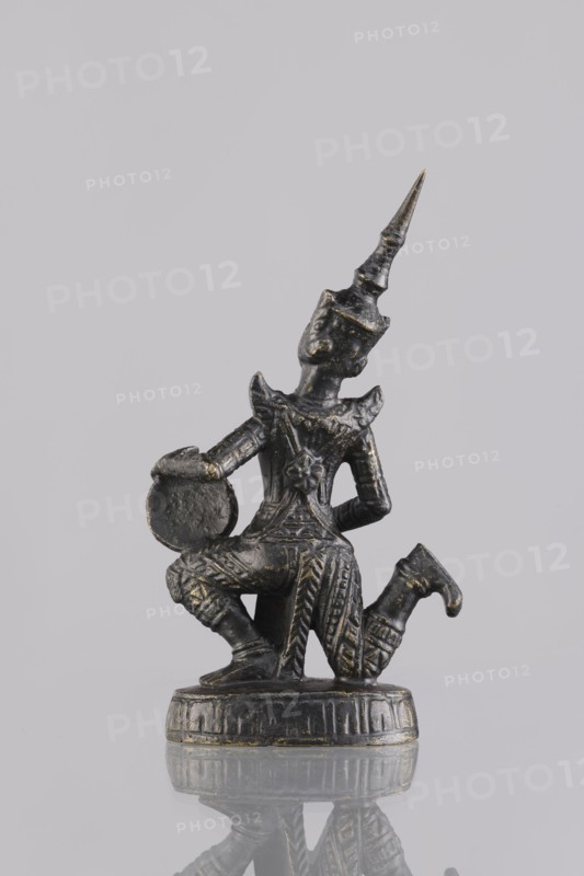 Musician figurine, Southeast Asian art, Southeast Asia culture, musicians, 1st half of the 20th century contemporary product, Cambodia, sculpture, figurine, height 10.8 cm, A small dark metallic figurine with traces of gold paint shows a kneeling figure wearing an ornate pointed headdress and richly patterned clothing; the figure holds a round object near one knee and rests on a circular decorated base, all set against a pale gray background with a reflection visible beneath the base.
