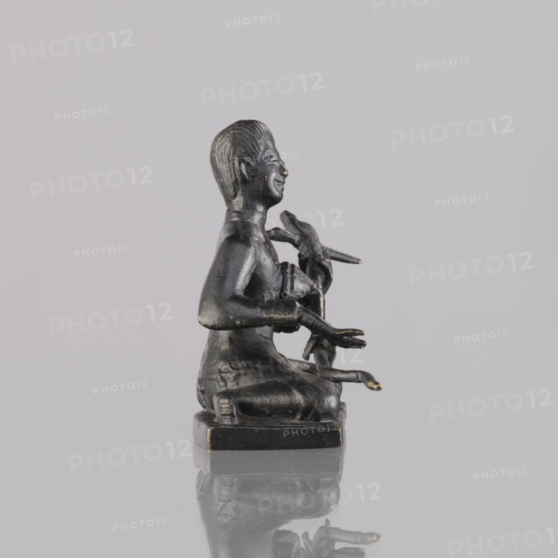 Musician figurine, art of Southeast Asia, Southeast Asia culture, musicians, 1st half of the 20th century contemporary product, Cambodia place of origin, sculpture, figurine, height 6.4 cm, A small dark bronze statuette of a seated human figure on a square base, shown in profile facing right, with textured hair and a visible smile; the figure holds an object with several elongated elements in its hands and arms, and there are subtle worn highlights of a lighter metallic tone on raised areas; the background and surface are light gray and the base casts a faint reflection on the glossy surface beneath.