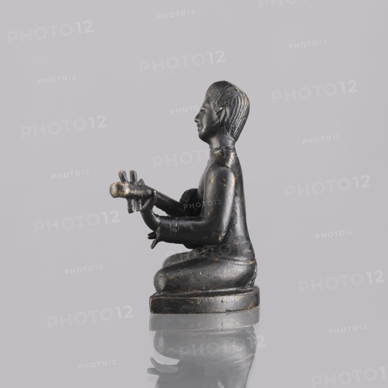 Musician figurine, art of Southeast Asia, Southeast Asia culture, musicians, 1st half of the 20th century contemporary product, Cambodia place of origin, sculpture, figurine, height 6.3 cm, A small dark statue of a seated human figure shown in profile, facing left, with hands extended forward holding a cylindrical object with a few protrusions; the figure has a smooth dark surface with patches of worn lighter tone, sculpted hair texture, and a rounded base that sits on a reflective surface; the background is a uniform pale gray, and the visible colors are dark black, muted gold or bronze wear, pale gray, and reflections of those tones.