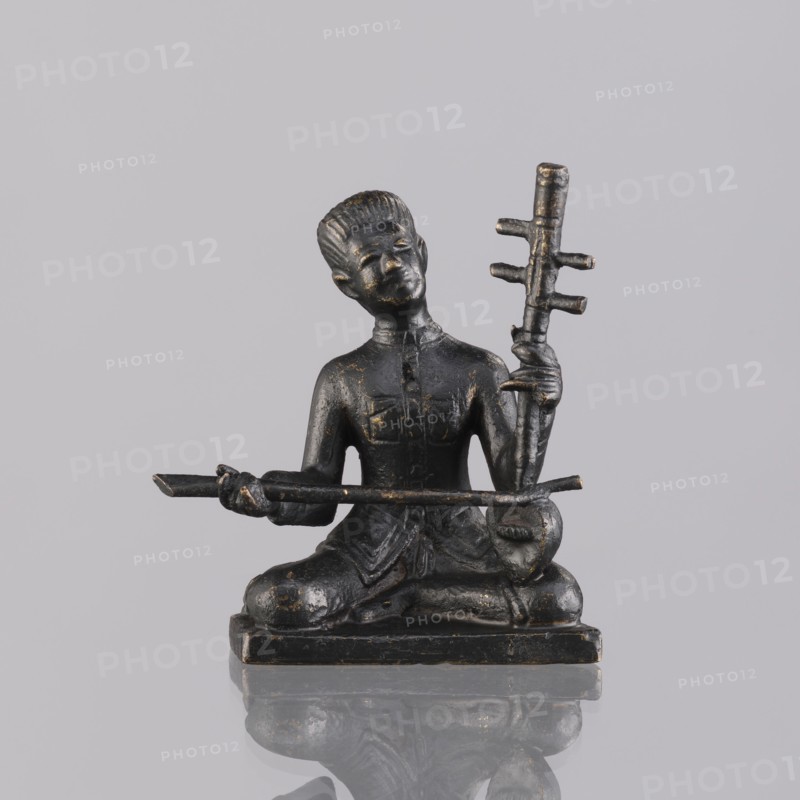 Musician figurine, Southeast Asian art; subject: musicians; culture: Southeast Asia, Cambodia; provenance, 1st half of the 20th century contemporary product; sculpture, figurine; height 6.3 cm, A small dark statue of a seated figure with a shiny surface and worn golden highlights, posed cross-legged on a rectangular base, holding a long horizontal rod with both hands and a vertical staff with several protrusions in one hand; the figure has a sculpted hairstyle and clothing details, and the whole object sits on a reflective pale gray surface against a pale gray background with visible reflections.