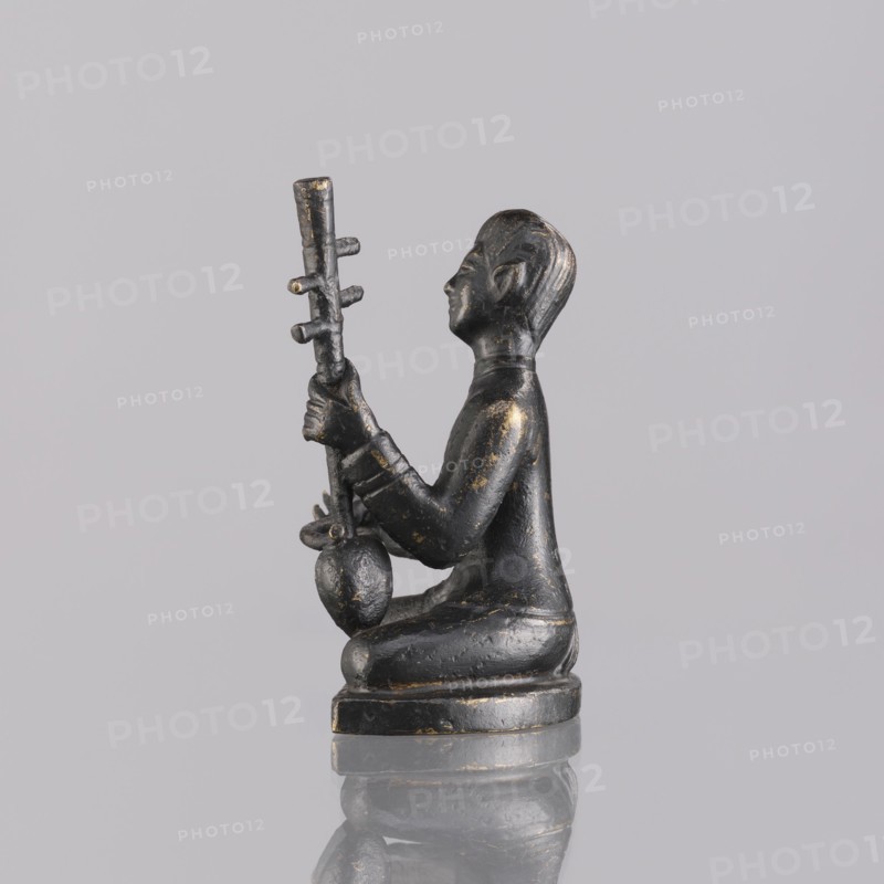 Musician figurine, Southeast Asian art, musicians, Southeast Asia culture, 1st half of the 20th century contemporary product, Cambodia, sculpture, figurine, height 6.3 cm, A small dark figurine sits on a rounded base, shown in profile facing right, with a smooth dark surface accented by worn golden highlights; the figure is seated with legs folded beneath, wearing a long-sleeved garment, and holding a vertical object with several short protrusions and a bulbous element near its lower end; the head has stylized hair and facial features with a serene expression; the figure and base rest on a reflective surface that mirrors their shapes against a pale gray background.