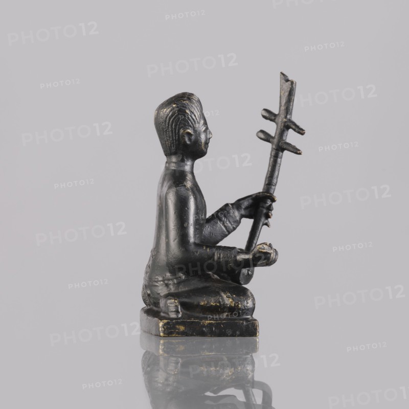 Musician figurine, art of Southeast Asia, Southeast Asia culture, musicians, 1st half of the 20th century contemporary product, Cambodia place of origin, sculpture, figurine, height 6.5 cm, A small dark metallic figurine of a seated person shown in profile, with a smooth dark surface and traces of worn gold highlights, sitting on a rectangular base; the figure holds a vertical staff with several horizontal protrusions, has short ridged hair, and is posed with bent arms and hands grasping the staff, all set against a pale light gray background with a subtle reflective surface beneath showing a faint mirrored outline.