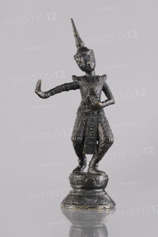 Dancer figurine, dance, Southeast Asian art, Southeast Asia culture, 1st half of the 20th century contemporary product, Cambodia place of origin, sculpture, figurine, height 13.2 cm, A small dark metallic statue of a human figure stands on a round ornate base, the figure wearing a tall pointed headdress and detailed patterned clothing, posed with one arm extended outward and the other bent toward the torso; the surface shows hints of worn metallic highlights. The overall colors visible are dark gray to black with touches of faded gold.