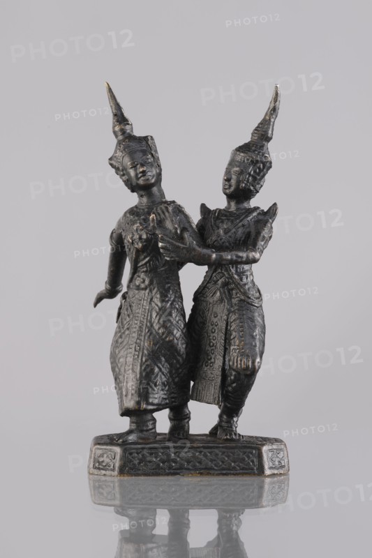 Figurines of a dancing couple, dance, Southeast Asia culture, art of Southeast Asia, 1st half of the 20th century, Cambodia, sculpture, figurine, height 11.7 cm, A small dark metallic statue of two standing figures on a rectangular base, both wearing tall pointed headdresses and long intricately patterned garments; one figure stands slightly forward with an arm extended back while the other stands close behind with an arm touching the first figure’s chest, their faces and hands visible with smooth sculpted features, and the base shows decorative carved patterns; visible colors include dark gray to black metal with subtle hints of bronze or brown tones, and a neutral light gray background.