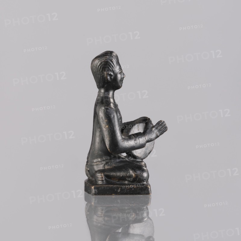 Musician figurine, art of Southeast Asia, Southeast Asia culture, musicians, 1st half of the 20th century contemporary product, Cambodia place of origin, sculpture, figurine, height 6.3 cm, A small dark statue of a seated figure viewed in profile, shown kneeling on a square base with hands raised and holding a rounded object, rendered with smooth surfaces and subtle carved hair texture, placed on a reflective surface; visible colors include black with hints of dark brown and a neutral gray background.