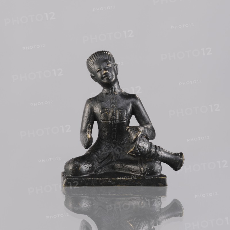 Musician figurine, Southeast Asian art; topic: musicians; culture: Southeast Asia; provenance, 1st half of the 20th century contemporary product; origin: Cambodia; sculpture, figurine; height 6.3 cm, A small dark figurine of a seated person with a glossy black surface and hints of worn gold highlighting, seated cross-legged on a rectangular base with a reflective surface beneath, the figure wearing a fitted upper garment with subtle carved details, one hand resting on the knee and the other holding a rounded object near the lap, the head tilted slightly and showing short textured hair and a calm facial expression, background a smooth light gray.