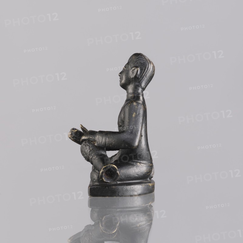 Musician figurine, Southeast Asian art, musicians, Southeast Asia culture, 1st half of the 20th century contemporary product, Cambodia, sculpture, figurine, height 6.3 cm, A small sculpted figure in a seated pose made of a dark material with worn golden highlights, shown in profile facing left, with a defined head, facial features, and molded hair, wearing a long-sleeved garment and seated on a rounded base; the figure holds a cylindrical object across its lap with visible texture and open ends, and the object and the figure show areas of wear revealing lighter tones; the scene rests on a reflective surface, and the background is a plain pale gray.