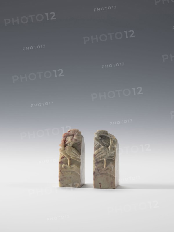 Pair of seal dies, Chinese, 19th–20th century. Materials/technique: blind, square cross-section; decoration of cranes in bas-relief and engraving; pendant form. Dimensions: height 6.9 cm, width 2.7 cm, depth 2.7 cm., A pair of carved stone objects stand side by side on a flat white surface against a smooth gradient gray background, each object rectangular with a rounded top and showing carved reliefs of birds and foliage; the stones display a mix of muted greens, beige, pale yellow, and touches of reddish-brown, with subtle variations and mottling across their surfaces, casting soft shadows on the surface beneath them.