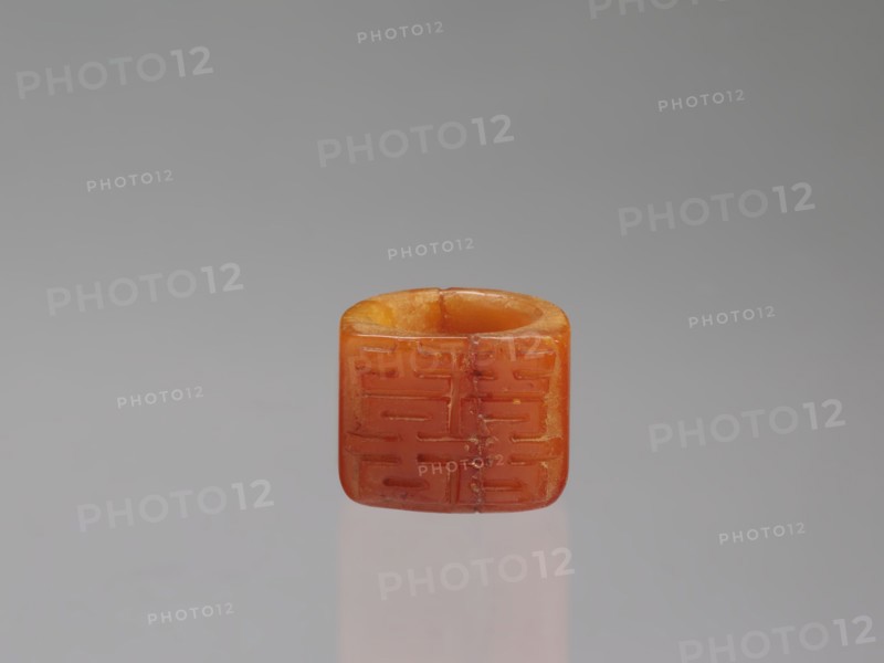 Signet ring, Chinese, Qing dynasty 1644–1911, opaque amber with convex relief, 18th–19th century, China; jewelry; ornament symbolizing happiness; height 3.3 cm, width 3.1 cm, depth 2.6 cm., An orange, square-shaped bead with rounded edges sits centered against a smooth gray background, showing a hollowed top and subtle surface grooves on its faces; the bead displays variations of orange and amber tones with small darker inclusions and a soft reflection beneath it on the surface.