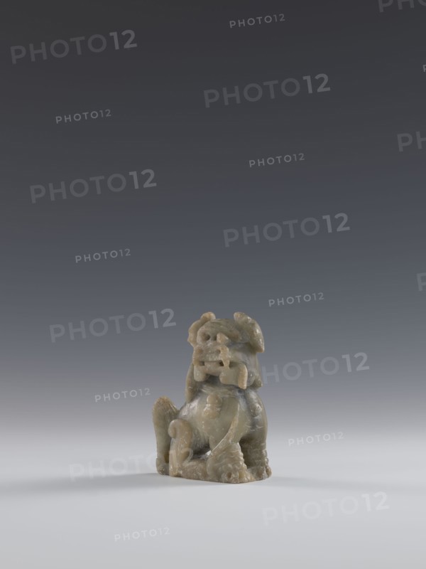 Seated lion, Chinese, lion fo iconography, Qing dynasty 1644–1911. Figurine carved in soapstone elephantine, engraved. 18th–19th century. Sculpture/figurine. Height 10.5 cm; width 7.5 cm; depth 4.0 cm., A small carved figurine made of a greenish-gray stone sits on a smooth light-gray surface against a dark gray gradient background; the figurine depicts a seated creature with a rounded body, curled tail, prominent snout, open mouth, stylized limbs, and textured surface, showing variations of green, gray, and subtle brown tones.