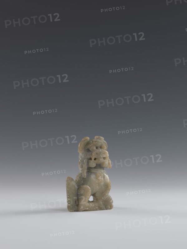 Seated lion, Chinese, lion fo iconography, Qing dynasty 1644–1911; soapstone elephantine carved and engraved figurine, 18th–19th century; sculpture/figurine; H 10.5 cm, W 7.5 cm, D 4.0 cm., A small carved figurine sits on a smooth surface against a gradient background. The figurine appears to be made of a muted greenish-beige stone with lighter and darker mottling. It has a rounded head with pronounced facial features and a prominent open mouth, flanked by curled elements that hang down beside the face. The body is compact with a curved back and folded limbs, and textured detailing suggests fur or scales along parts of the form. The surface shows subtle variations in sheen and some pitted or weathered areas. The scene includes colors of gray in the background, white in the foreground surface, and greenish-beige in the figurine.