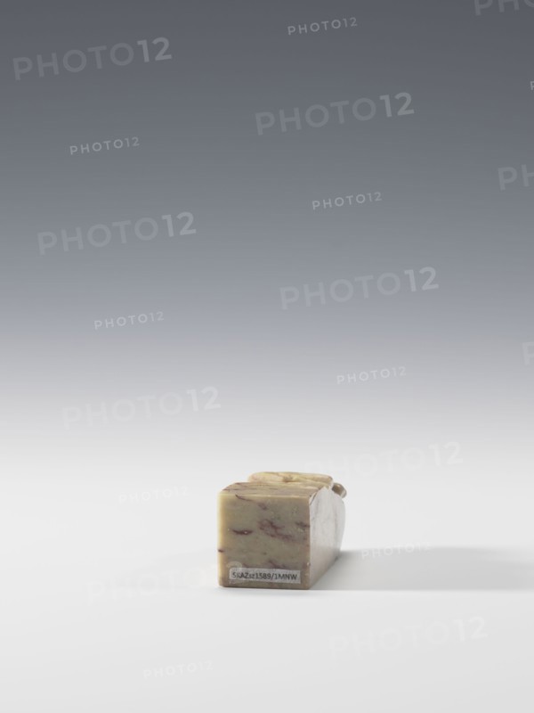 Seal stamp, Chinese, Qing dynasty 1644–1911, cranes iconography. Seal stamp, blind, square cross-section. Decoration: cranes bas-relief, engraving. Pendant, 19th–20th century, China. Dimensions: height 6.9 cm; width 2.7 cm; depth 2.7 cm., A small rectangular block of solid material rests on a flat surface near the lower center of the image, showing a marbled pattern with muted greenish-beige tones and streaks of reddish-brown; the block has slightly rounded top edges and appears smooth with a faint shadow extending to the right, set against a background that transitions from white at the bottom to a gradient of gray toward the top.