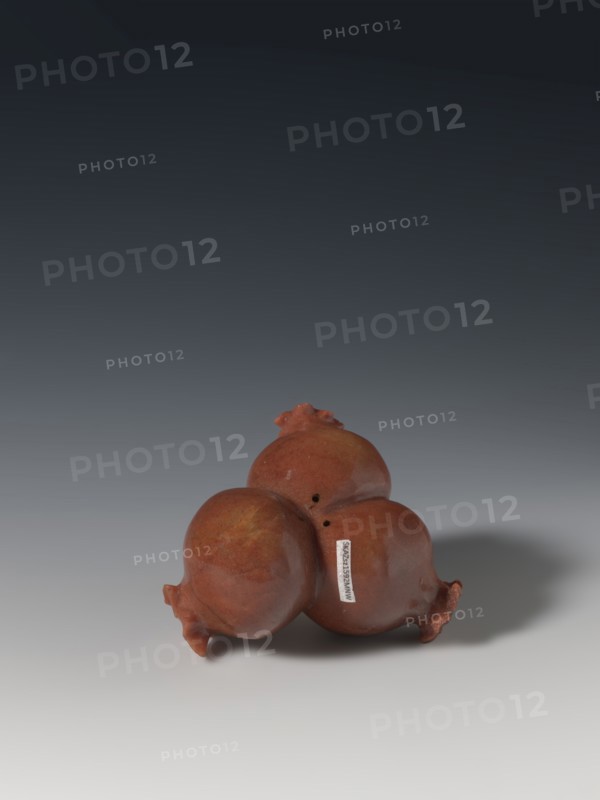 Three connected pomegranates fragment of a whole., Chinese; Qing dynasty 1644–1911. Object in the shape of three connected pomegranates, each with small holes on both sides; fragment of a whole. 18th–19th century. Sculpture; material/varia. Height 4.8 cm, width 8.2 cm. Pomegranates fruits; pendants., A group of three round, pomegranate-shaped objects clustered together, each with a rough textured surface and small crown-like protrusions; the objects are reddish-brown with areas of darker and lighter brown and a subtle glossy sheen. They sit on a smooth surface that transitions from light gray near the foreground to darker gray toward the background, and a soft shadow falls to the right of the cluster. The background above the surface fades from medium gray to a deep nearly black gradient.